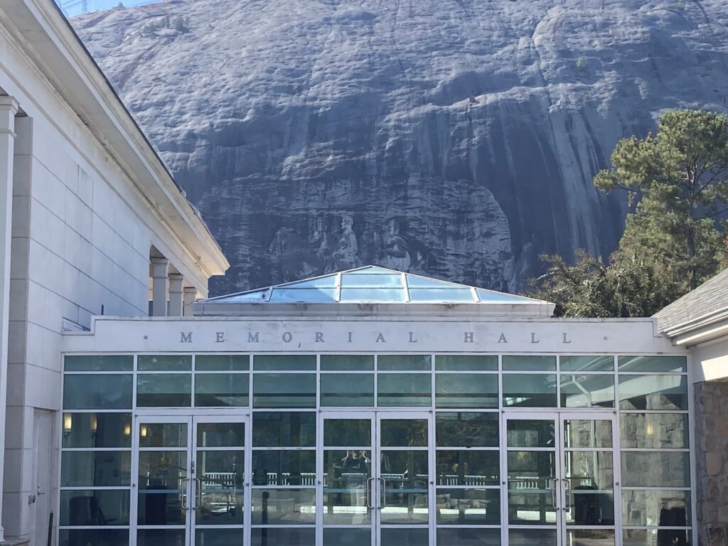 A photo of the Stone Mountain carving, the lower half of the image covered by the 'Memorial Hall' in the foreground of the image.